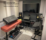 A studio setting showing various music equipment including a Moog Sub 37 synthesizer under a dust cover, along with other synthesizers and audio equipment.